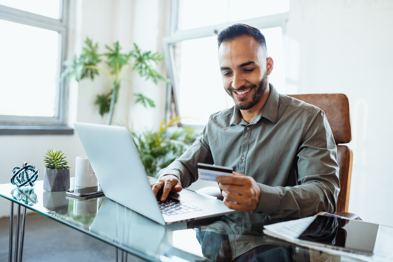 Empresário sorrindo enquanto usa um cartão de crédito empresarial em frente ao notebook, em um escritório moderno com plantas e itens de trabalho sobre a mesa.
